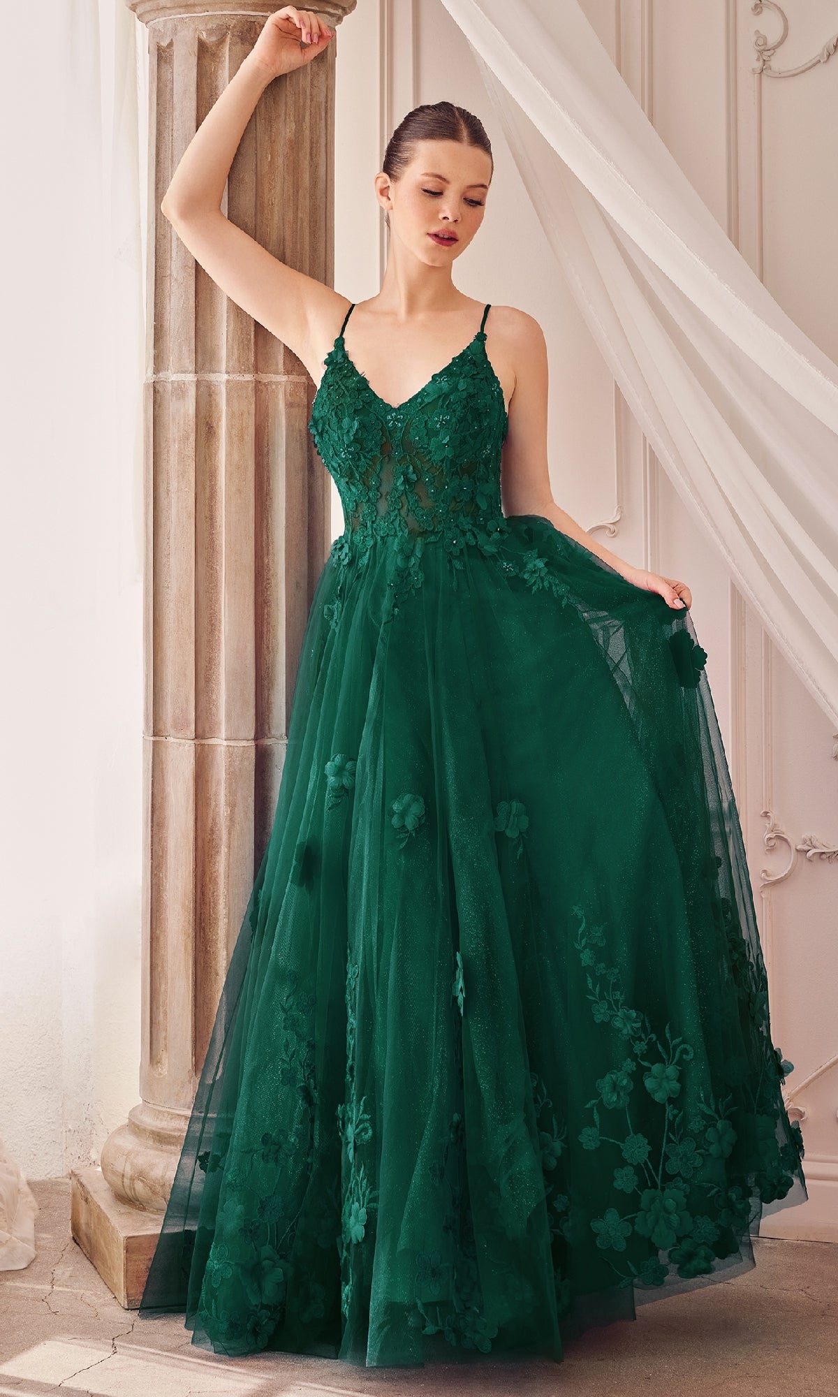 A woman poses indoors by a stone column, holding part of her skirt while wearing the Andrea and Leo A1326 Long Glitter Prom Ball Gown, a sleeveless, floor-length formal dress with floral detailing.